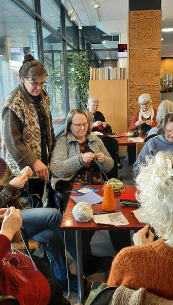 Käsityökahvilassa voi jutella tai istua neulomassa hiljaa itsekseenkin, kahvilan pöydistä kerrotaan. Anna-Liisa Heinonen seisomassa taustalla.